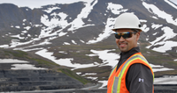 Smiling worker in safety gear stands in front of snowy, mountainous landscape at an industrial site.